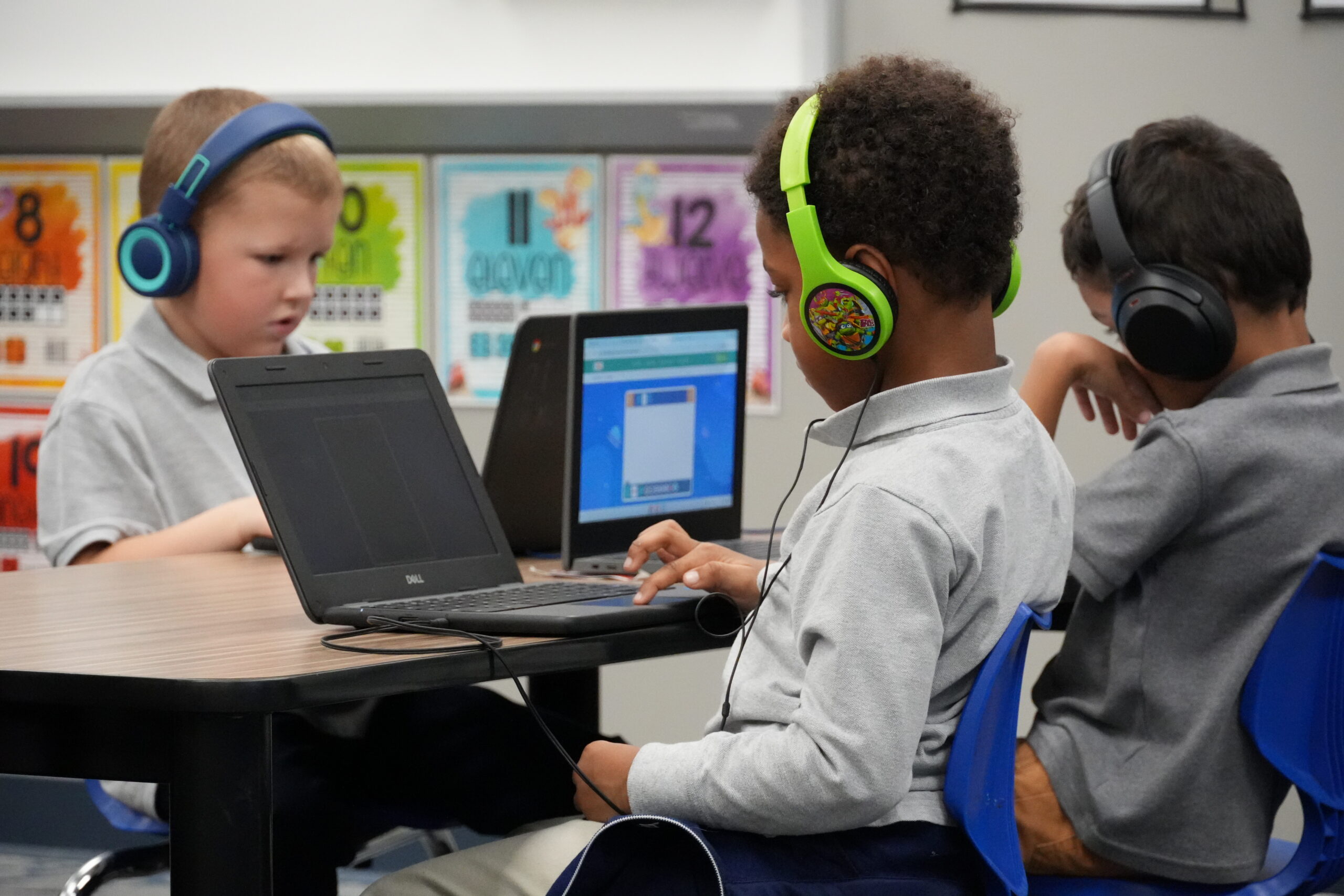 Three students wearing headphones use laptops during a classroom learning activity.