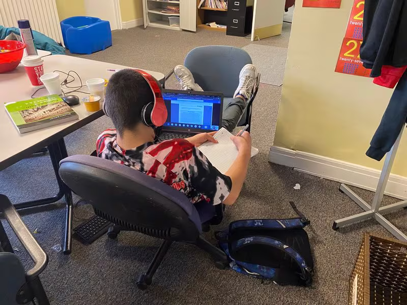 ZTN3VVRRG5CYDCDBKL5VYIVV7U Student wearing headphones works independently on a laptop while taking notes.