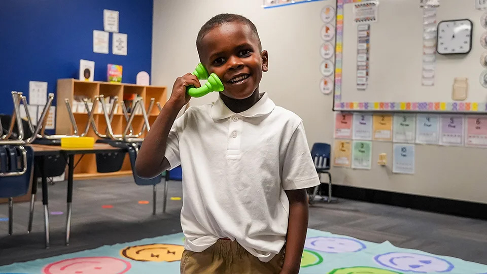 ajb-features-3-2 Smiling Andrew J. Brown Academy student holding a green toy telephone during a classroom learning activity