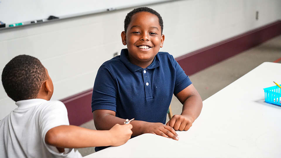 Smiling Andrew J. Brown Academy student sitting at a classroom table with a classmate during a learning activity