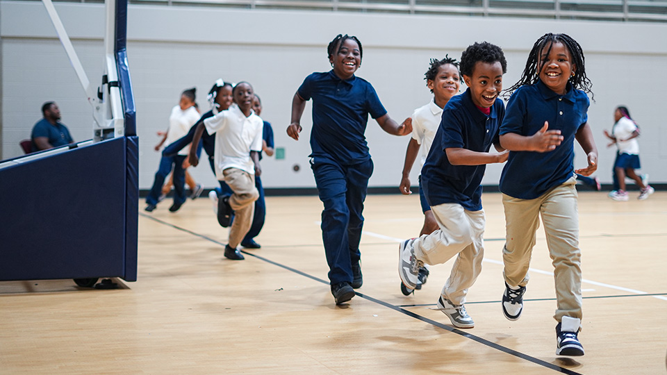 Andrew J. Brown Academy students smiling and running together during an indoor physical education activity in the school gymnasium