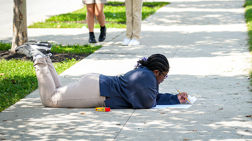 Paramount Englewood student lying on the sidewalk while writing on a worksheet outdoors