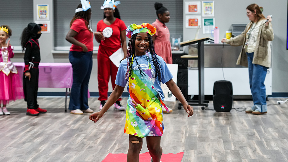 Girls IN STEM Academy student smiling and walking in a colorful costume during a school celebration, with classmates and staff in the background
