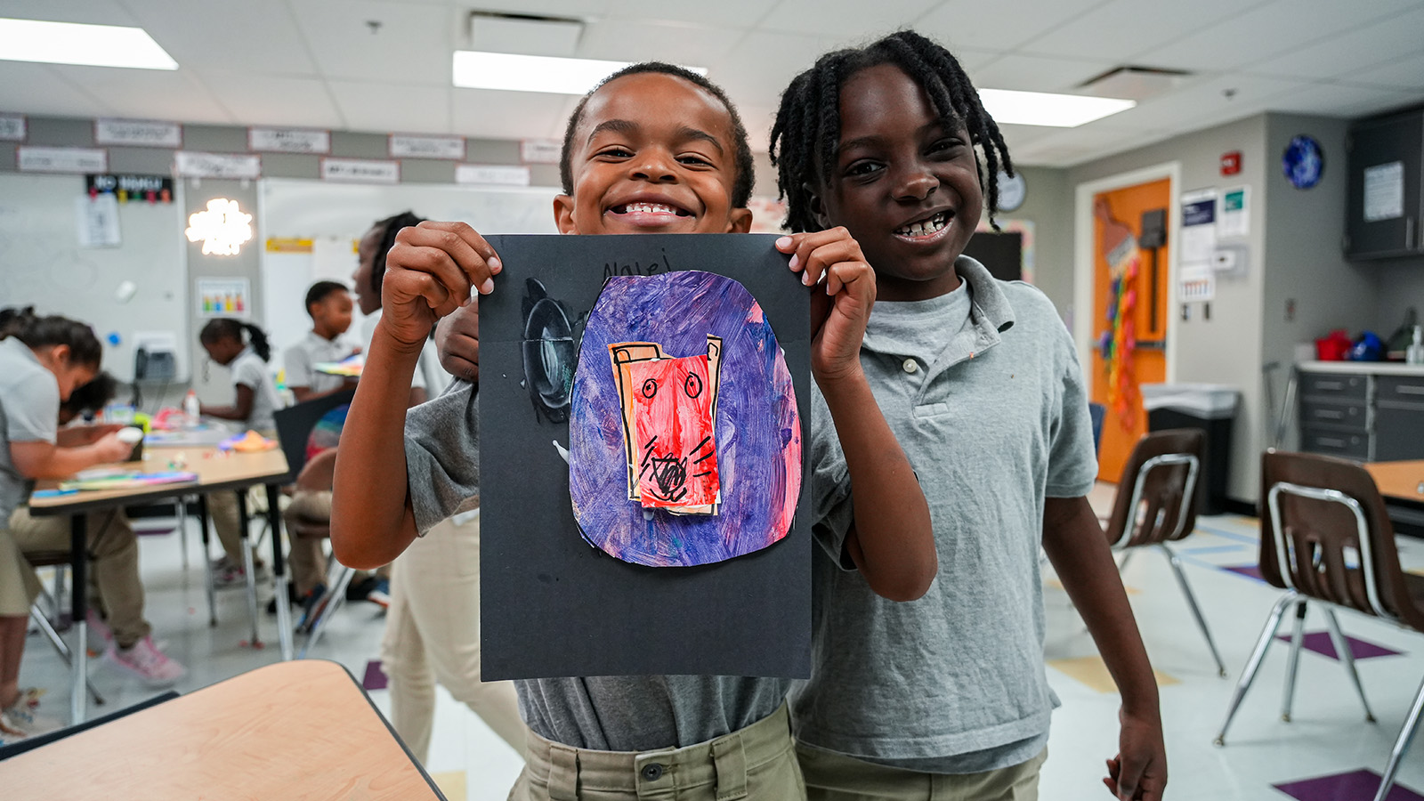Two smiling Paramount Lafayette students proudly displaying their colorful artwork during a classroom art activity