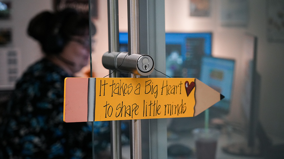 Decorative pencil-shaped sign at Paramount Online Academy reading It takes a big heart to shape little minds, with a teacher working in the background