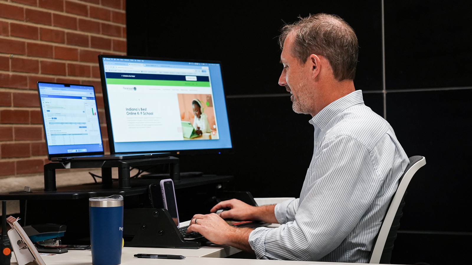 Paramount Online Academy Principal Anthony Brose working at his desk with a laptop and computer monitors in his office
