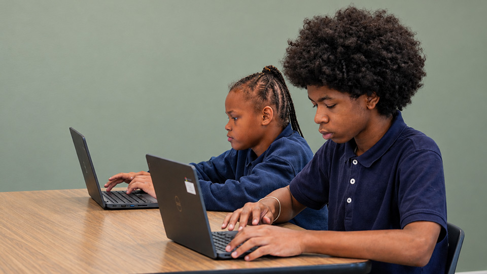 Two Paramount Online Academy students working independently on laptops while completing school assignments side by side.