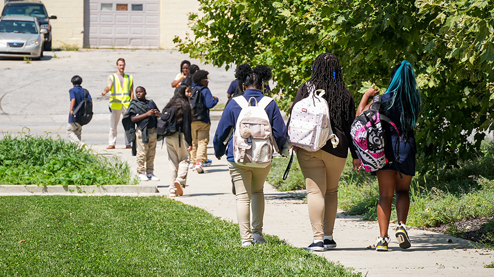 Paramount students walking along a sidewalk with backpacks as school staff supervise their arrival or dismissal